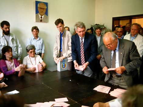 Ein Mann wertet einen Stimmzettel aus. Um ihn herum sind viele Menschen, die ihn beobachten.