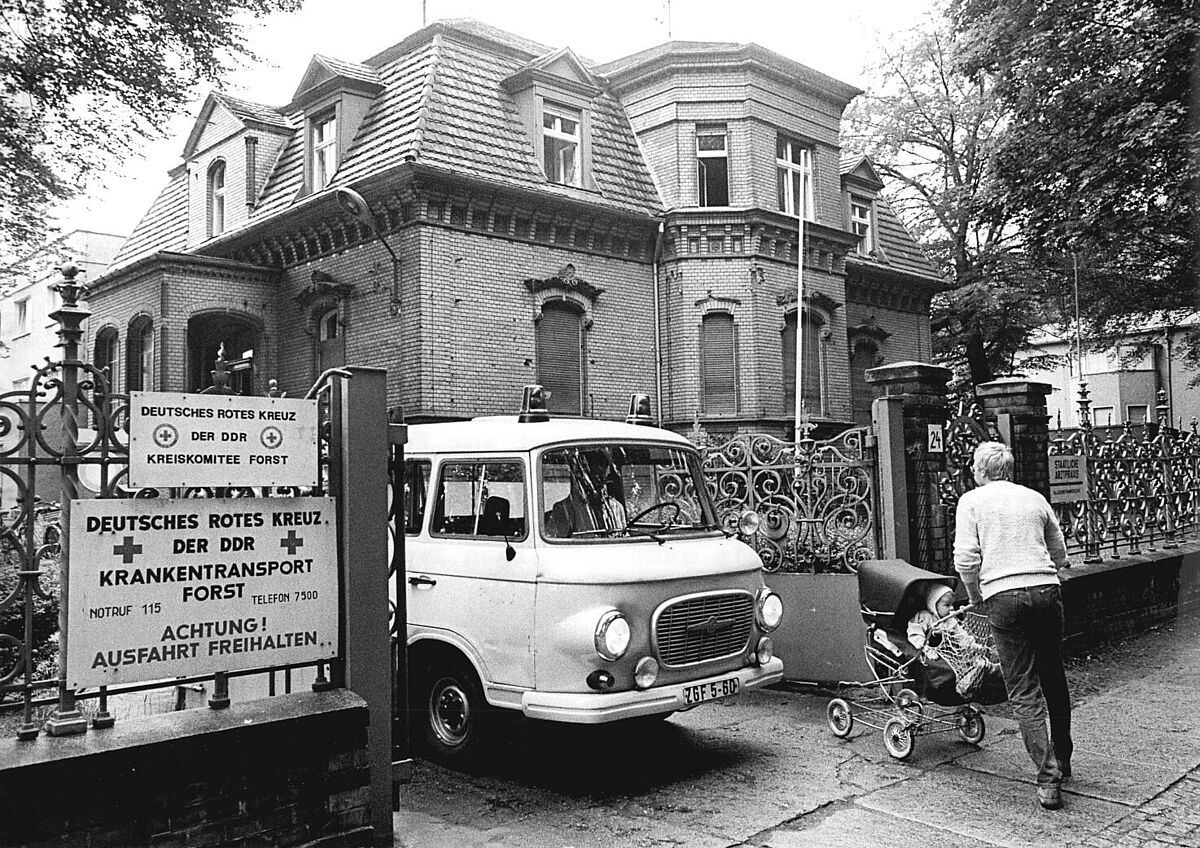 Ein Auto des Krankentransports fährt durch das Tor des Deutschen Roten Kreuz in Forst, während ein Mann mit Kinderwagen seitlich vorbeigeht. Im Hintergrund ist die gemauerte Villa zu sehen, in der sich die DRK-Stelle befindet.