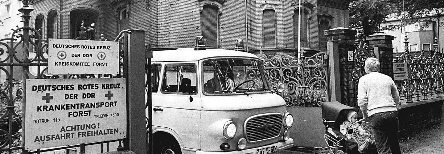 Ein Auto des Krankentransports fährt durch das Tor des Deutschen Roten Kreuz in Forst, während ein Mann mit Kinderwagen seitlich vorbeigeht. Im Hintergrund ist die gemauerte Villa zu sehen, in der sich die DRK-Stelle befindet.