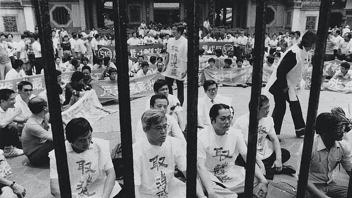 Auf dem Schwarz-Weiß-Bild zu sehen sind männliche auf dem Boden sitzende Demonstranten in heller Kleidung.