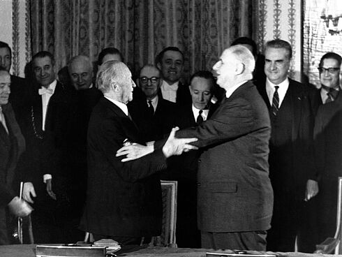 Konrad Adenauer und Charles de Gaulle strecken ihre Arme zueinander aus, um sich zu umarmen. Im Hintergrund stehen mehrere Menschen, die lächeln.
