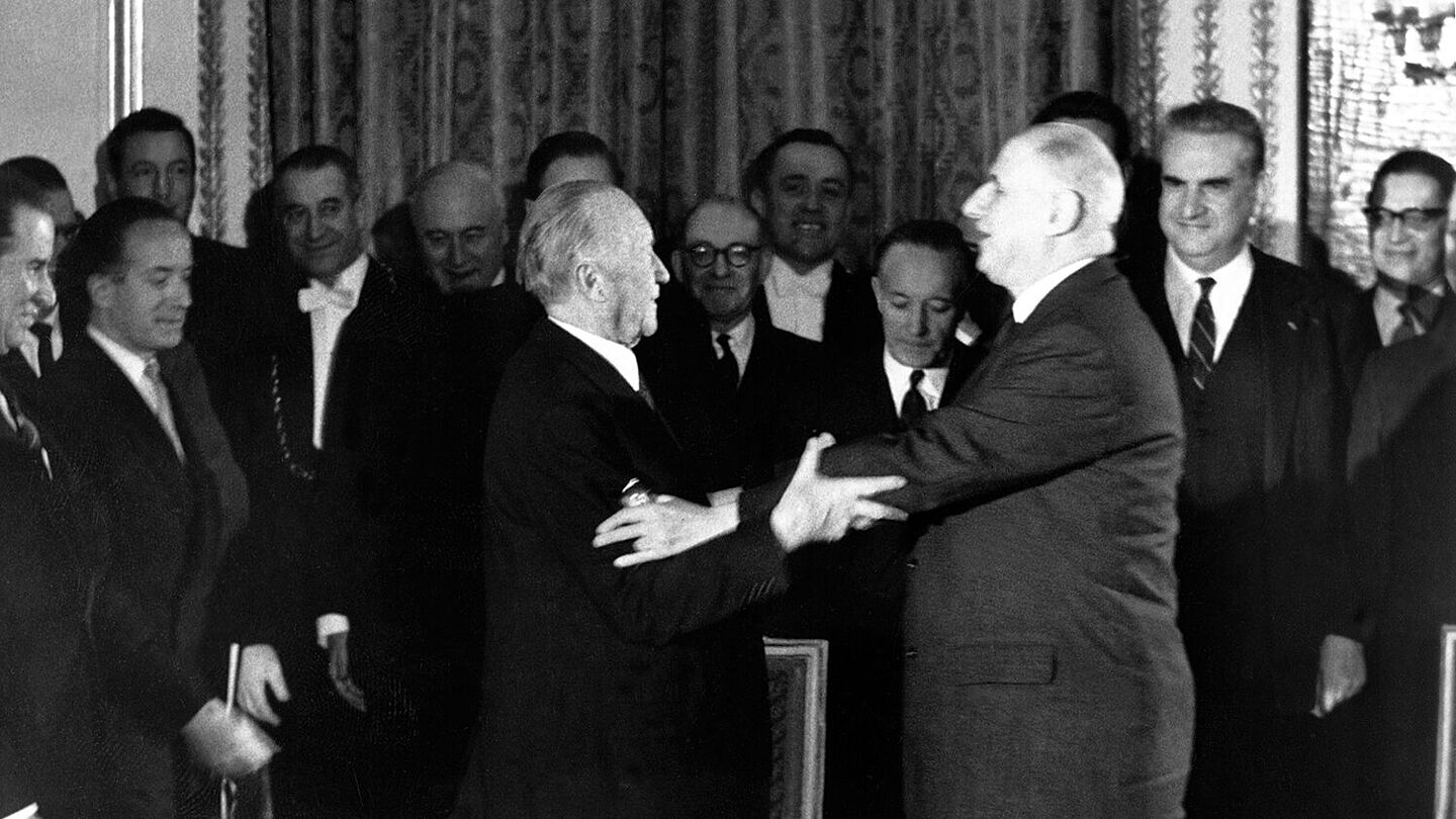 Konrad Adenauer und Charles de Gaulle strecken ihre Arme zueinander aus, um sich zu umarmen. Im Hintergrund stehen mehrere Menschen, die lächeln., Quelle:
            BArch, B 145 Bild-00361399 / Schwahn, Ernst
