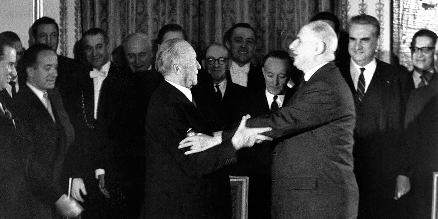 Konrad Adenauer und Charles de Gaulle strecken ihre Arme zueinander aus, um sich zu umarmen. Im Hintergrund stehen mehrere Menschen, die lächeln., Quelle:
            BArch, B 145 Bild-00361399 / Schwahn, Ernst