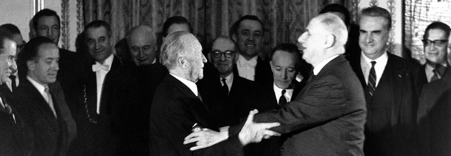Konrad Adenauer und Charles de Gaulle strecken ihre Arme zueinander aus, um sich zu umarmen. Im Hintergrund stehen mehrere Menschen, die lächeln.