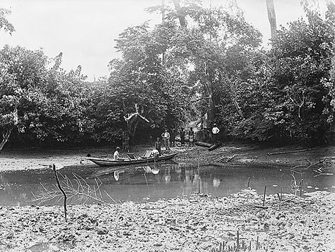 Die Schwarz-Weiß-Aufnahme des Fotografen Carl Hohl zeigt einen schmalen Wasserlauf. Mehrere Personen stehen am bewaldeten Ufer und sehen dabei zu, wie ein Mann an einem Kahn hantiert. Ein weiterer Mann sitzt in dem Boot.