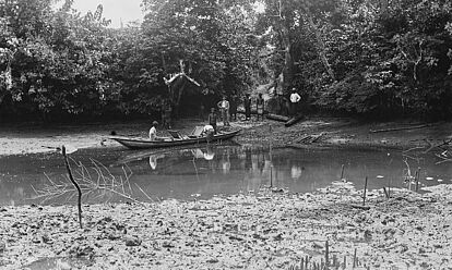 Die Schwarz-Weiß-Aufnahme des Fotografen Carl Hohl zeigt einen schmalen Wasserlauf. Mehrere Personen stehen am bewaldeten Ufer und sehen dabei zu, wie ein Mann an einem Kahn hantiert. Ein weiterer Mann sitzt in dem Boot.