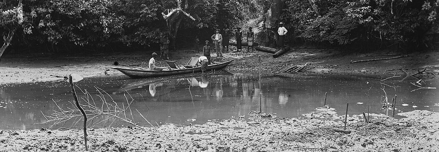 Bootsfahrer auf einem afrikanischen Fluss vor Mangroven Die Schwarz-Weiß-Aufnahme des Fotografen Carl Hohl zeigt einen schmalen Wasserlauf. Mehrere Personen stehen am bewaldeten Ufer und sehen dabei zu, wie ein Mann an einem Kahn hantiert. Ein weiterer Mann sitzt in dem Boot.