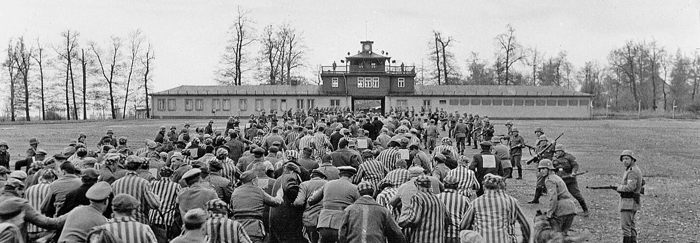 Schwarz-weiß-Bild: eine dem Betrachtenden abgewandte Gruppe Menschen, teilweise in Häftlingskleidung wird von bewaffneten Soldaten in Richtung eines Tores getrieben.