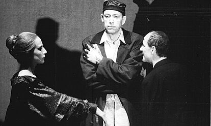 Drei Personen stehen auf der Bühne. In der Mitte, den Betrachtenden frontal zugewandt, die Arme auf der Brust verschränkt, mit neutralem Gesichtsausdruck Jörg Gudzuhn als Claudius. Links neben ihm, in Seitenansicht Dagmar Manzel als Gertrud, den rechten ausgestreckten Arm auf Brusthöhe angehoben, das Gesicht wirkt starr. Als dritte Person ganz rechts Ulrich Mühe als Hamlet, der mittigen Person zugewandt, so dass er eher von hinten zu sehen ist. Er scheint aber zu sprechen.