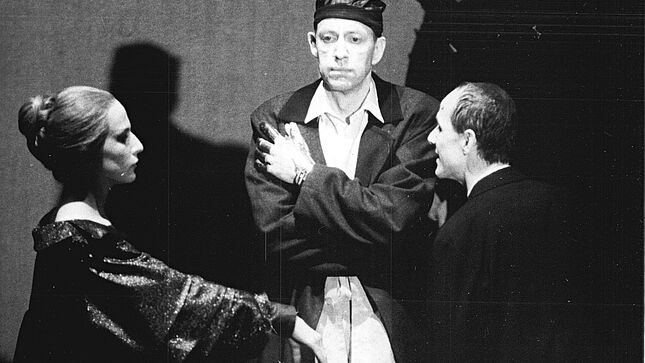 Drei Personen stehen auf der Bühne. In der Mitte, den Betrachtenden frontal zugewandt, die Arme auf der Brust verschränkt, mit neutralem Gesichtsausdruck Jörg Gudzuhn als Claudius. Links neben ihm, in Seitenansicht Dagmar Manzel als Gertrud, den rechten ausgestreckten Arm auf Brusthöhe angehoben, das Gesicht wirkt starr. Als dritte Person ganz rechts Ulrich Mühe als Hamlet, der mittigen Person zugewandt, so dass er eher von hinten zu sehen ist. Er scheint aber zu sprechen.