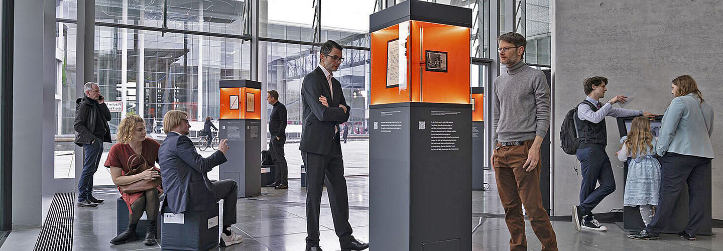 Ausstellungsstelen verteilt in einem Raum mit Glasfront, Menschen betrachten die ausgestellten Objekte.