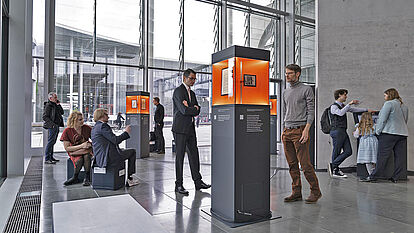 Ausstellungsstelen verteilt in einem Raum mit Glasfront, Menschen betrachten die ausgestellten Objekte.