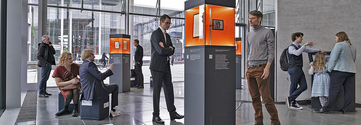 Ausstellungsstelen verteilt in einem Raum mit Glasfront, Menschen betrachten die ausgestellten Objekte.