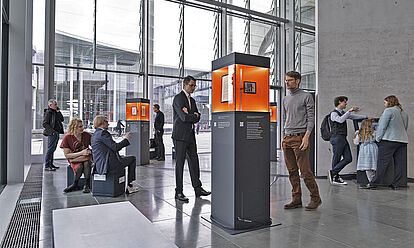 Ausstellungsstelen verteilt in einem Raum mit Glasfront, Menschen betrachten die ausgestellten Objekte.