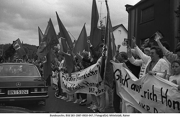 Bei der Fahrt der Auto-Kolonne von Erich Honecker durch Wiebelskirchen säumen zahlreiche Schaulustige die Strecke. DKP-Mitglieder begrüßen den Staatsratsvorsitzenden mit roten Fahnen und Plakaten, 10. September 1987