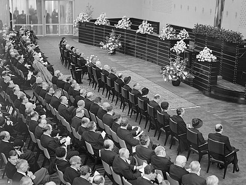 Schwarz-Weiß-Aufnahme mehrerer Stuhlreihen, die besetzt sind mit Personen in dunkler, festlicher Trauerkleidung, in Richtung eines mit großen Blumengestecken geschmückten Podests blickend.