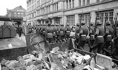 In Reihe marschierende NVA-Soldaten ziehen vorbei an einem Schuttcontainer im Vordergrund, in dem auch ein Portrait von Erich Honecker liegt. Die Soldaten sind Teil des Wachregiments „Friedrich Engels“ und auf dem Weg zur Neuen Wache in Berlin.