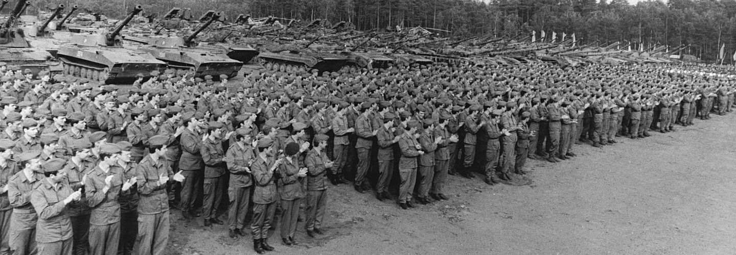 Mehrere Reihen angetretener Soldaten der Landstreitkräfte der NVA applaudieren anlässlich eines Truppenbesuchs von Erich Honecker im Juni 1984. Im Hintergrund stehen mehrere Reihen gepanzerter Fahrzeuge und Hubschrauber.