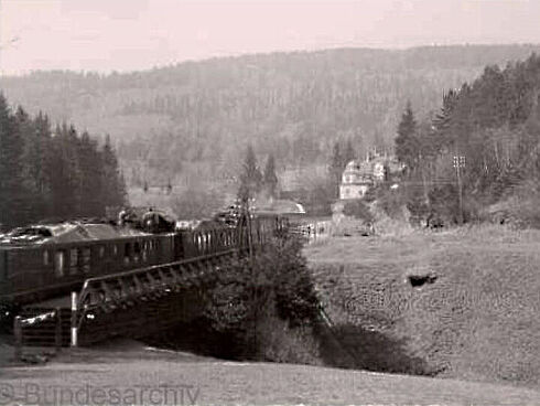 Schwarz-Weiß-Aufnahme eines Zuges, der inmitten einer hügeligen, bewaldeten Landschaft über eine Talbrücke fährt