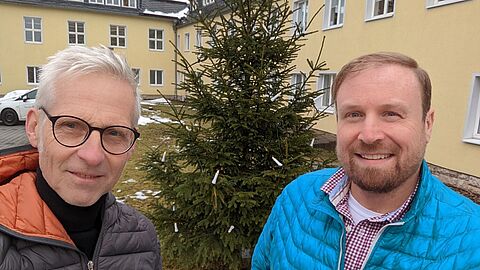 Maximilian Schönherr und Sascha Münzel stehen vor einem Weihnachtsbaum, der mit Lichtern geschmückt ist.