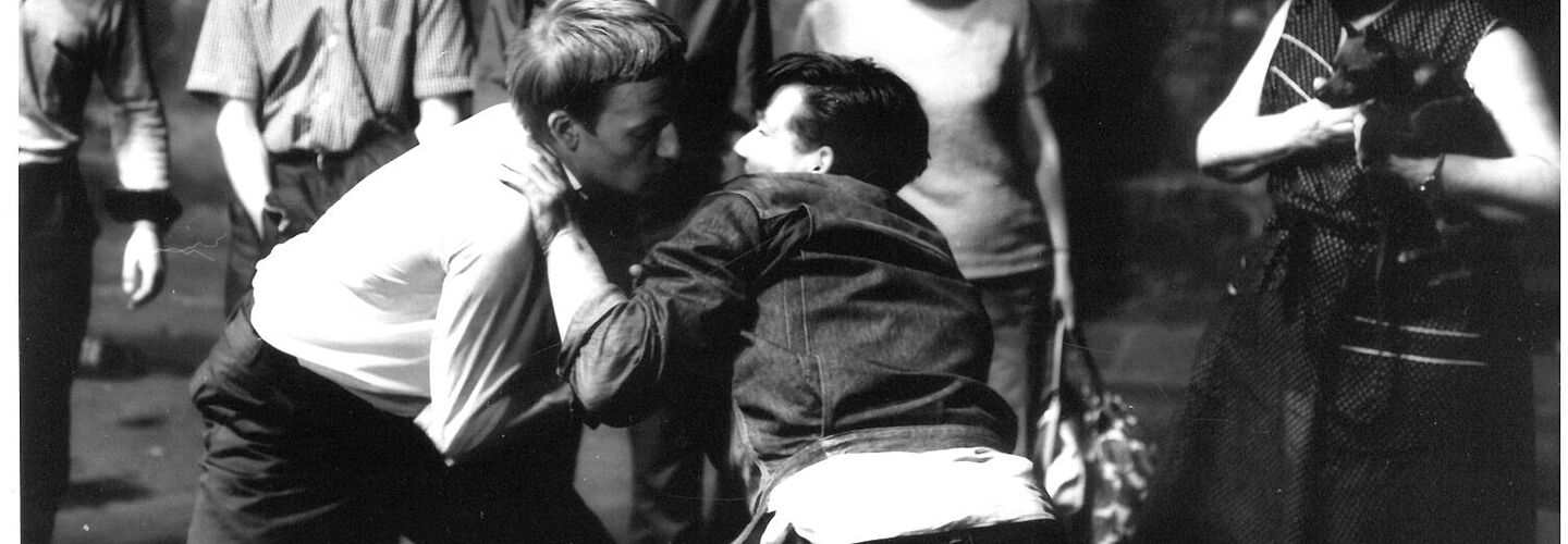 Zwei Jugendliche ringen miteinander, umstehende Personen schauen dem Kampf zu.