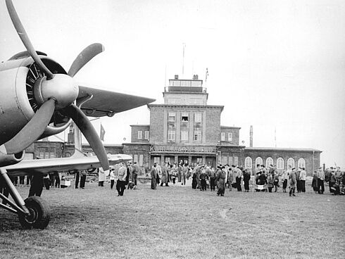 Im Vordergrund erkennt man den Propeller eines Flugzeuges. Im Hintergrund ist ein großes Flughafengebäude zu sehen, vor dem eine große Menschenmenge steht.