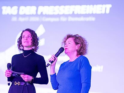 Tag der Pressefreiheit 2026 Zwei Personen mit Mikrofonen stehen auf einer Bühne. Im Hintergrund ist ein lila Hintergrund mit der Aufschrift „Tag der Pressefreiheit" zu sehen.