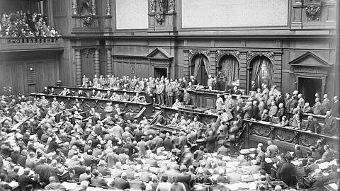 Schwarz-weiße Aufnahme eines vollen Plenarsaals im Reichstag mit Redner am Rednerpult und zahlreichen sitzenden und stehenden Abgeordneten