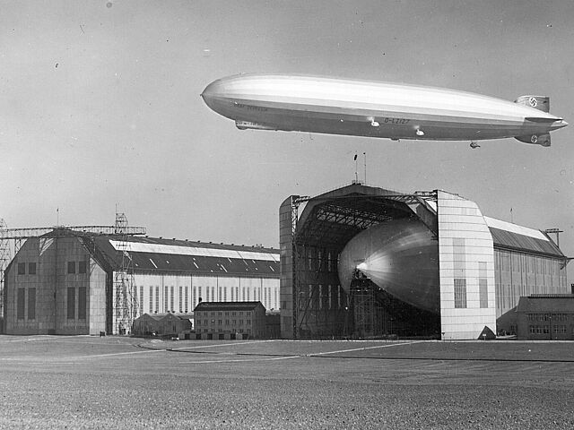 Das Luftschiff LZ 129 „Hindenburg“ während des Baus in der Luftschiffhalle in Friedrichshafen. Über der Halle fliegt das Luftschiff LZ 127 „Graf Zeppelin“.