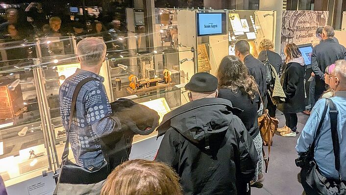 Rund 150 Gäste besuchten die Ausstellungseröffnung in Berlin-Lichterfelde Mehrere Personen schauen sich beleuchtete Vitrinen an, die mit Dokumenten gefüllt sind.