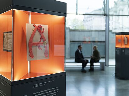 Eine Glasvitrine, in der sich ein aus Alufolie und Krepppapier gebasteltes "A" befindet.