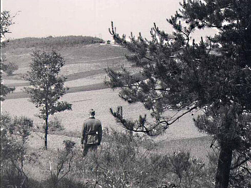 Auf dem Schwarz-Weiß-Foto ist ein Soldat zu sehen, der mit dem Rücken zum Fotografen auf einem mit Bäumen, Sträuchern und Gras bewachsenen Hügel steht und in die Richtung einer Feldlandschaft blickt. Auf einem der Hügel in der Ferne sind Zäune und Gebäude zu erkennen.