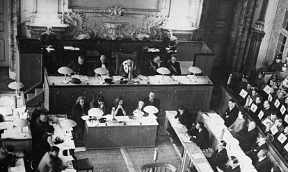 Schwarz-Weiß-Fo­to: Blick von schräg oben, gerade zu drei hinterewinander stehende Podien, das erste schmal mit 5 Personen, zwei Mikrofonen und zwei Tischlampen; dahinter mehr als doppelt so breit mit seitlicher Treppe mit 5 Personen, einem Mikrofon und 4 Tischlampen, dahinter noch etwas breiter mit jeweils einer außen sitzenden Person. Recht befinden sich zwei lange hintereinander stehende Tische mit jeweil 4 und 5 Personen. Dahinter eine Art Sitzrang mit mehreren Reihen Personen mit Zahlen vor der Brust. Links drei Reihen mit verschiedenen Schreibtischen und 7 Personen. Auf allen Tischen liegen viele Papiere.