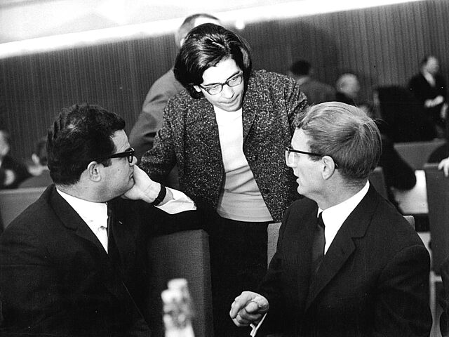 Schwarz-Weiß-Fotografie mit zwei sitzenden Herren und einer stehenden Frau bei einem Gespräch