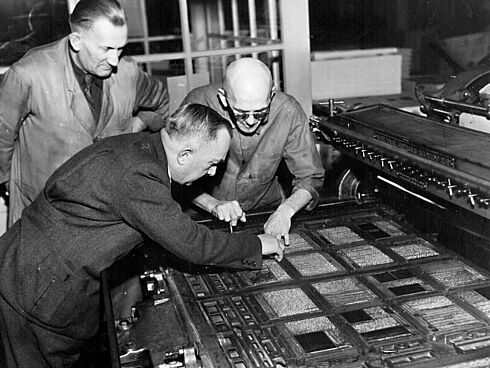 Druckerei des Verlages Schwarz-Weiß Fotografie der Druckerei des Verlages mit drei Männern gebeugt über eine Druckerpresse.