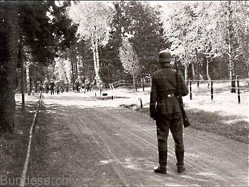 Im Vordergrund des Schwarz-Weiß-Bildes ist die Rückansicht eines uniformierten und mit einem Gewehr bewaffneten Soldaten zu sehen. Der Soldat steht auf einem von Bäumen gesäumten Sandweg und blickt in die Richtung einer Personengruppe, die sich auf ihn zu bewegt.