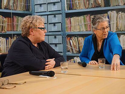 Evelyn Zupke​​​​​​​ und Ulrike Poppe im Gespräch mit einer Person außerhalb des Fotos. Im Hintergrund sieht man eine Wand, die vollständig mit einer Fototapete mit Regalen voller Stasiakten beklebt ist.