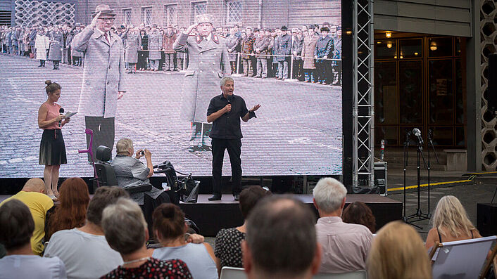 Im Hintergrund spricht Roland Jahn zu einem im Vordergrund befindlichen Publikum. Er steht vor einem großen Screen im Hintergrund, auf dem Erich Honecker und Erich Mielke vor Haus 1 zu sehen sind.