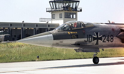 Ein Jagdflugzeug F-104G „Starfighter“ des Jagdgeschwaders 74 „Mölders“ steht auf einer Rollbahn. Im Hintergrund ist ein Tower zu sehen.