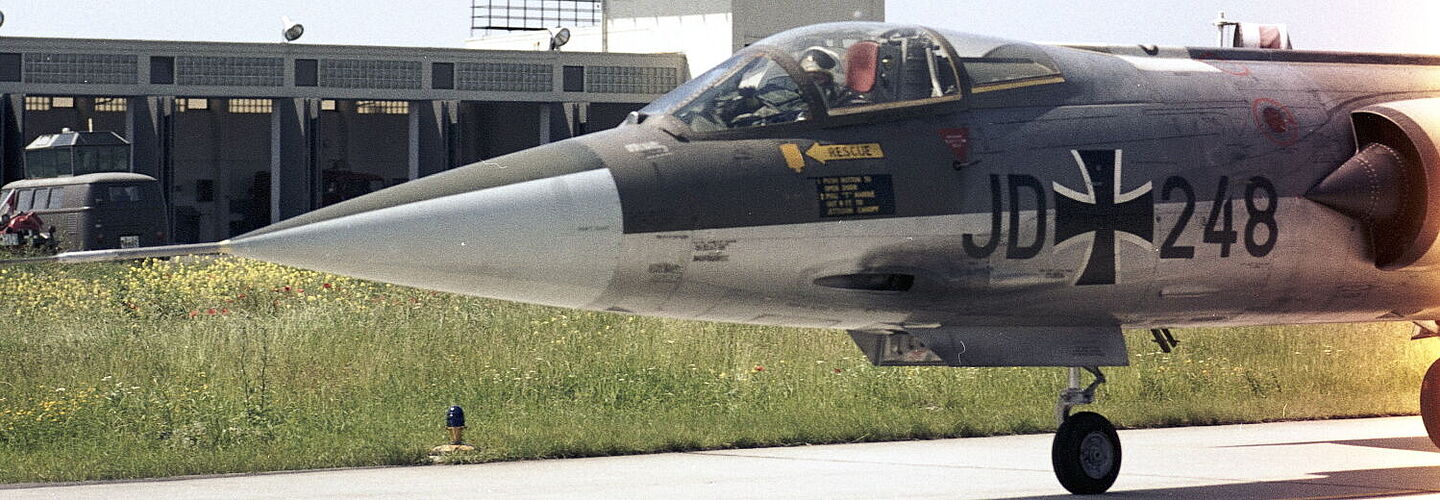 Ein Jagdflugzeug F-104G „Starfighter“ des Jagdgeschwaders 74 „Mölders“ steht auf einer Rollbahn. Im Hintergrund ist ein Tower zu sehen.