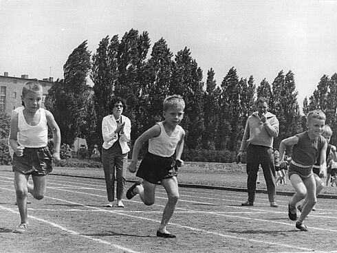 Mehrere Jungen in Sportkleidung laufen auf einer Rennbahn, während sie von einer Frau angefeuert werden.
