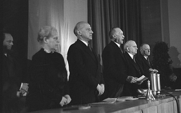 Konrad Adenauer steht mit einem Zettel in der Hand neben mehreren Personen und spricht.