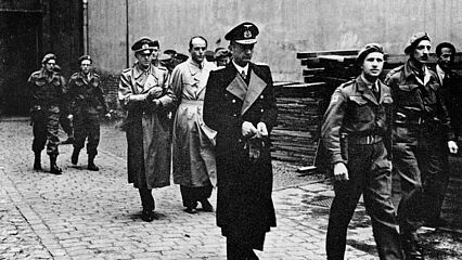 Schwarz-Weiß-Aufnahme mit drei Herren in Trenchcoats, die von uniformierten Soldaten begleitet werden.