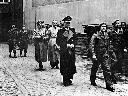 Schwarz-Weiß-Aufnahme mit drei Herren in Trenchcoats, die von uniformierten Soldaten begleitet werden.