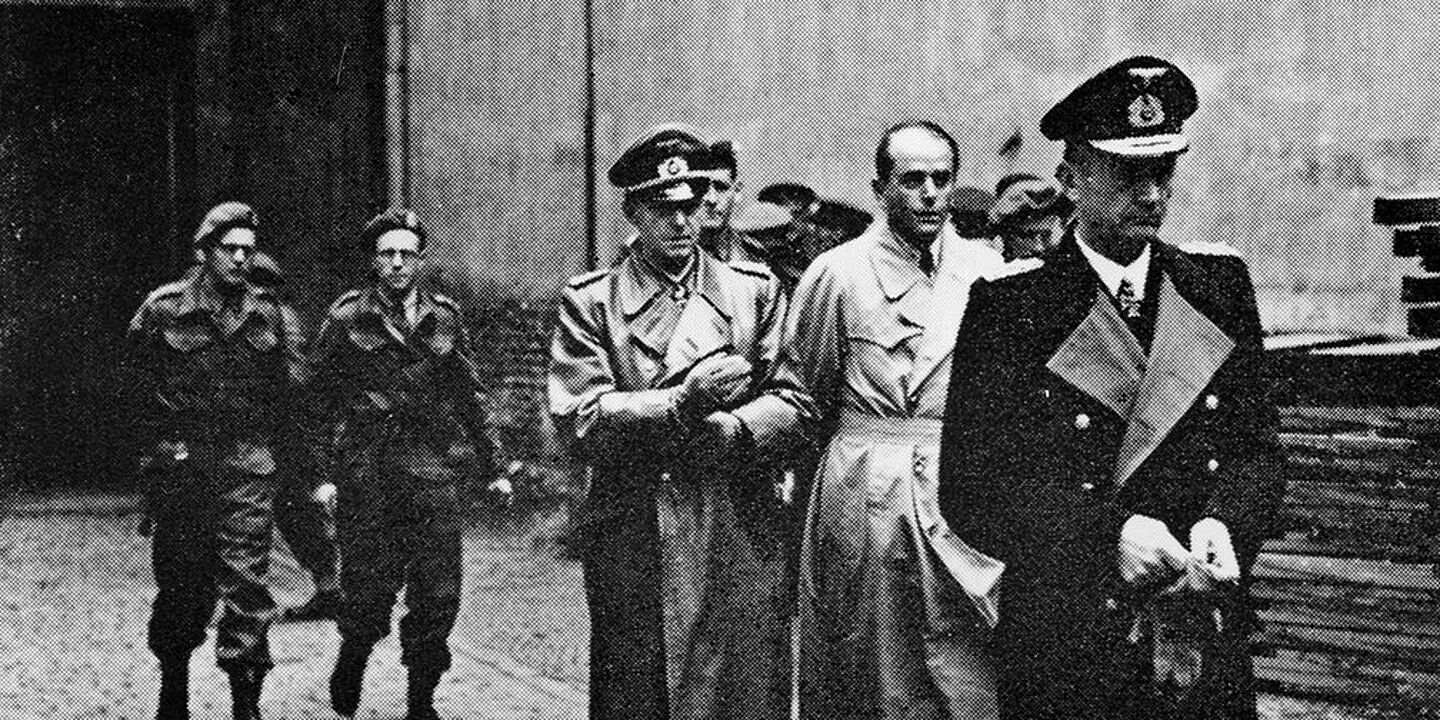 Verhaftung der Regierung Dönitz Schwarz-Weiß-Aufnahme mit drei Herren in Trenchcoats, die von uniformierten Soldaten begleitet werden., Quelle:
BArch, Bild 146-1985-079-31 / o. Ang.