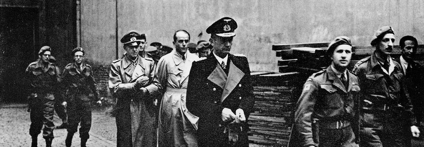 Schwarz-Weiß-Aufnahme mit drei Herren in Trenchcoats, die von uniformierten Soldaten begleitet werden.