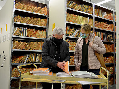 Das Bild zeigt den Bundesbeauftragten Roland Jahn und die Bundestagsabgeordnete Melanie Bernstein vor einem kleinen Transportwagen. Darauf liegen mehrere Stasi-Akten, in die beiden hineinschauen.