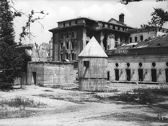 Von Bombentreffern beschädigtes Gebäude, vor dem sich der Eingang zum „Führerbunker“ befindet. Daneben steht ein kegelförmiger Bombenunterstand mit spitzem Dach.