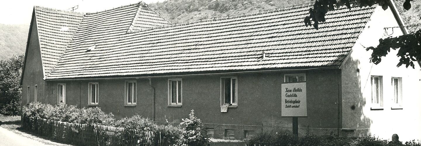 Ein Haus mit Hecke. Das Haus hat acht Fenster an der Front. Aus einem hängt ein Wäscheteil heraus. Ein Schild mit Aufschritt ist zu erkennen. Im Hintergrund ein Wald.
