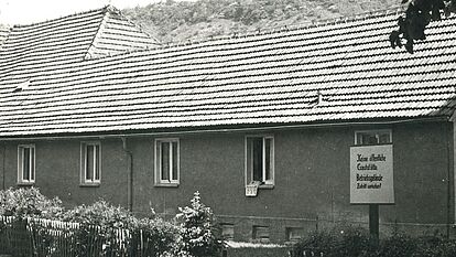 Ein Haus mit Hecke. Das Haus hat acht Fenster an der Front. Aus einem hängt ein Wäscheteil heraus. Ein Schild mit Aufschritt ist zu erkennen. Im Hintergrund ein Wald.